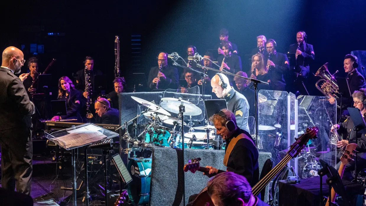 Muzikanten van het Orkest Koninklijke Luchtmacht spelen tijdens een concert, met in het midden fel verlicht de drummer.