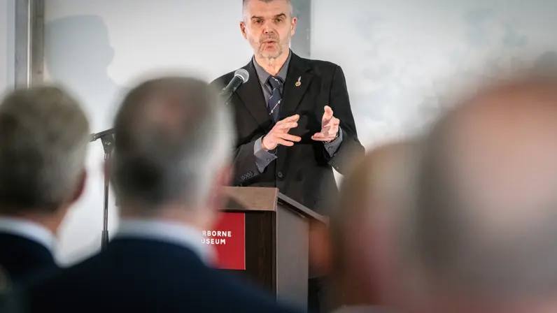 Een man met kort, grijs haar staat achter een spreekgestoelte. Hij draagt een donker pak. Op de voorgrond zijn achterhoofden te zien uit het publiek.&nbsp;