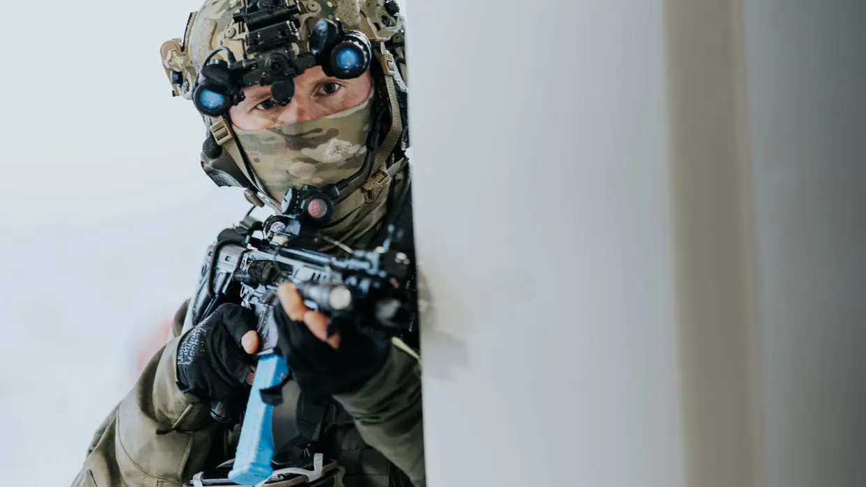 Links staan 2 NLMARSOF-operators  op de voorgrond, op de achtergrond zijn snelle onderscheppingsvaartuigen te zien die achter de veerboot varen. Rechts kijkt een gemaskerde NLMARSOF-operator om een hoekje, wapen gereed.
