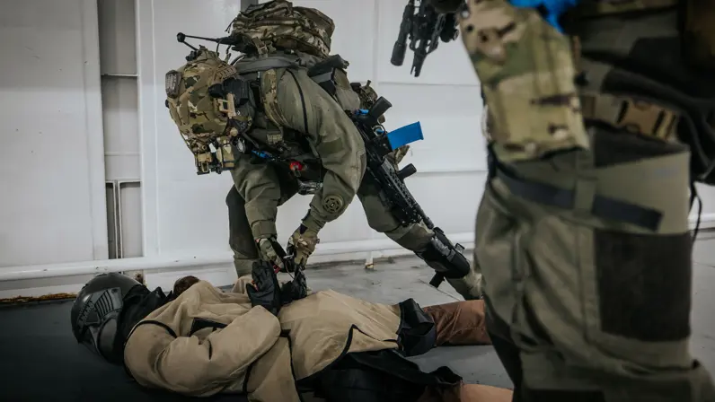 Een NLMARSOF-operator boeit een op de grond liggende oefenvijand. Een collega-operator kijkt toe. De oefenvijand draagt een helm en beschermende kleding. 