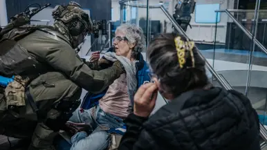 Links controleert een NLMARSOF-militair een vrouw die bleek ziet en duidelijk in nood verkeert. Rechts ondersteunen 2 gewapende NLMARSOF-operators een gewonde dame. Ze lopen over een scheepsdek.