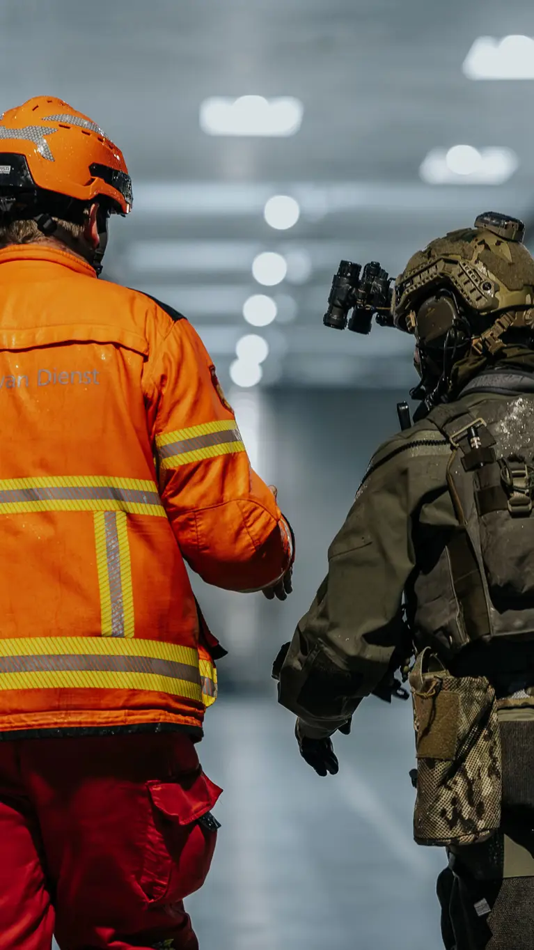 Alt: Twee figuren op de rug gezien, lopend door een verlicht scheepsruim. De linker heeft een fel oranje pak aan en helm op, de rechter is militair en draagt legergroen. 