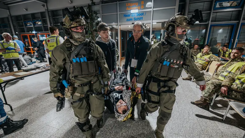Mariniers tillen met enkele mannen een gewond persoon. Zij komen binnen in een hal vol mensen en hulpverleners. 