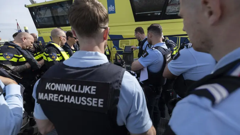 Marechaussees en politiemensen praten met elkaar op het dek van een schip en kijken naar de uitleg op een beeldscherm.