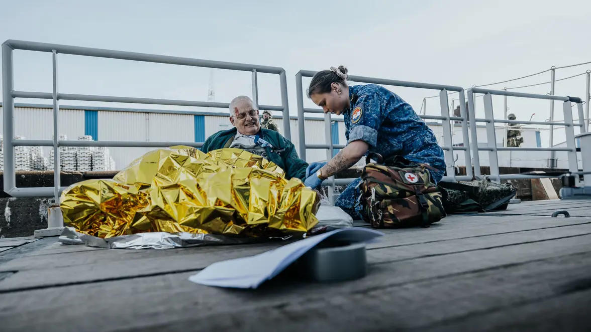 Een militair in een blauw vlekkenpak behandelt een gewonde burger op het dek van het schip. Over de man is een goudkleurige warmtedeken gedrapeerd.  