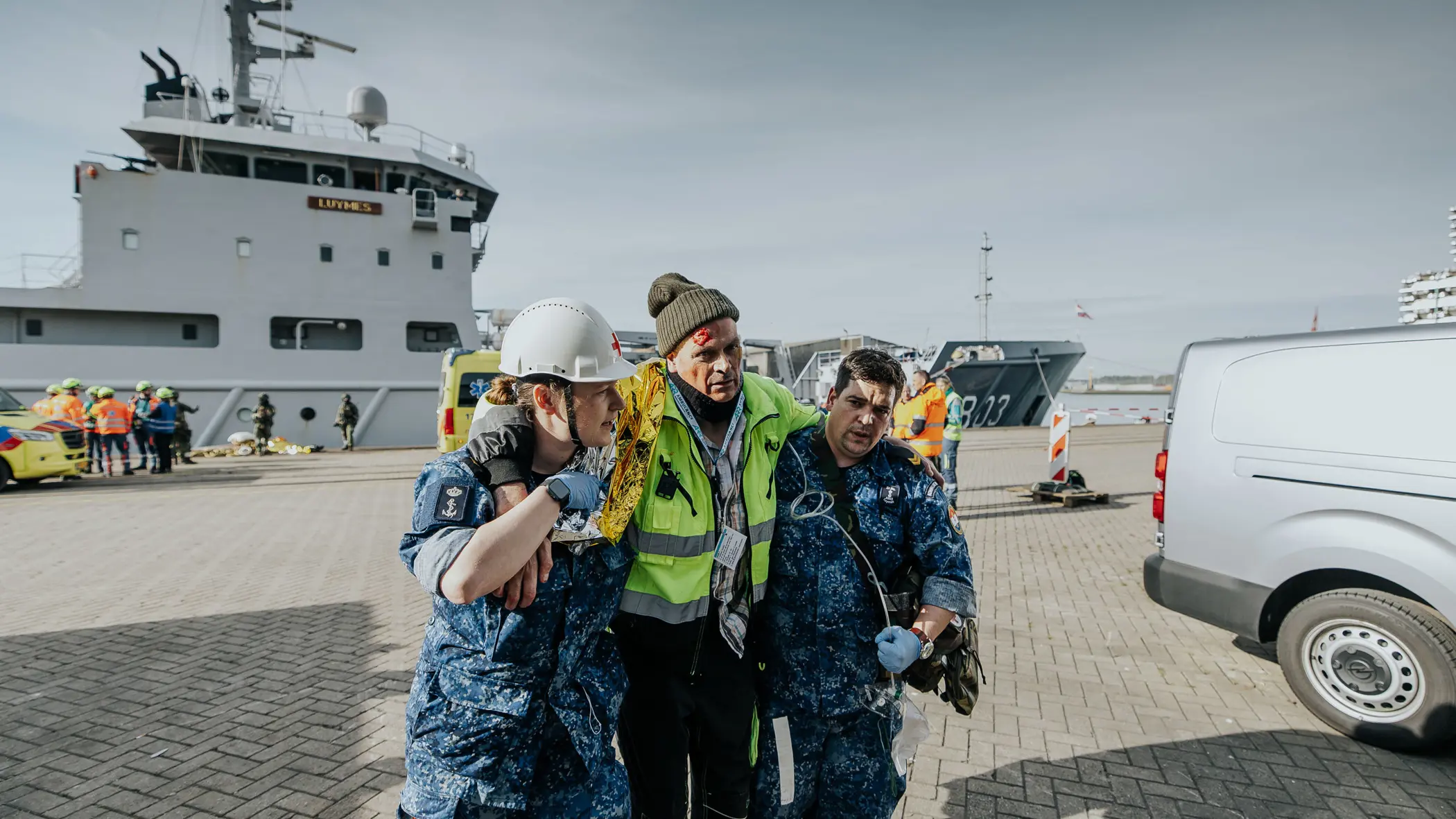 Een gewonde man wordt bij het lopen ondersteund door 2 militairen in boordtenue. Op de achtergrond is Zr.Ms. Luymes te zien.  