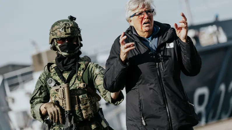 Een militair in een groen vlekkenpak begeleidt een gewonde vrouw. Ze schreeuwt en zwaait met haar handen.&nbsp;