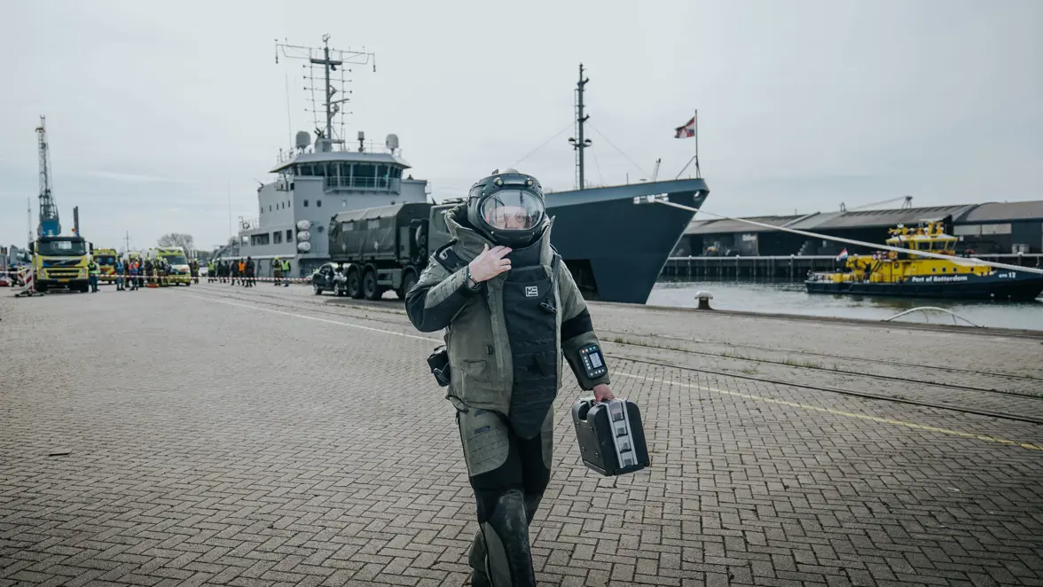 Een man in bompak loop over de kade. In zijn linkerhand draagt hij een kleine zwarte koffer. Achter hem is een schip te zien. 