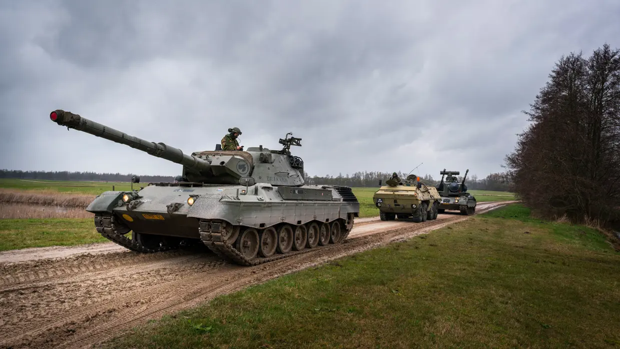 Een tank en gepantserde voertuigen rijden over een onverhard pad door een groen, open veld onder een bewolkte lucht. Rechts: Een zwaar militair voertuig met dubbele kanonnen en radar rijdt over een onverhard pad, gevolgd door andere legervoertuigen in een groen veld.