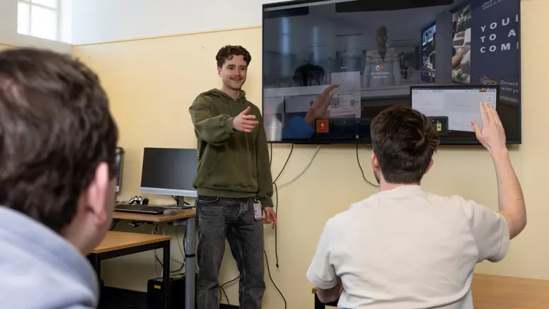 Een begeleider in een groene trui staat voor een digitaal schoolbord. Hij geeft de beurt aan een stagiair die zijn hand opsteekt.