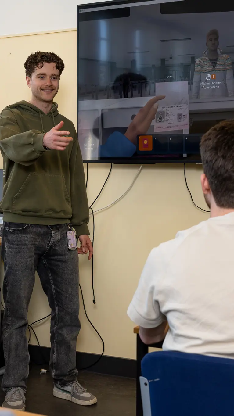 Een begeleider in een groene trui staat voor een digitaal schoolbord. Hij geeft de beurt aan een stagiair die zijn hand opsteekt.