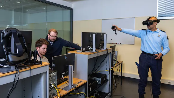 Twee mannen turen naar een computerscherm. Rechts staat een marechaussee die een VR-bril draagt. Hij heeft controllers in zijn handen en wijst met zijn rechterhand in de lucht. 