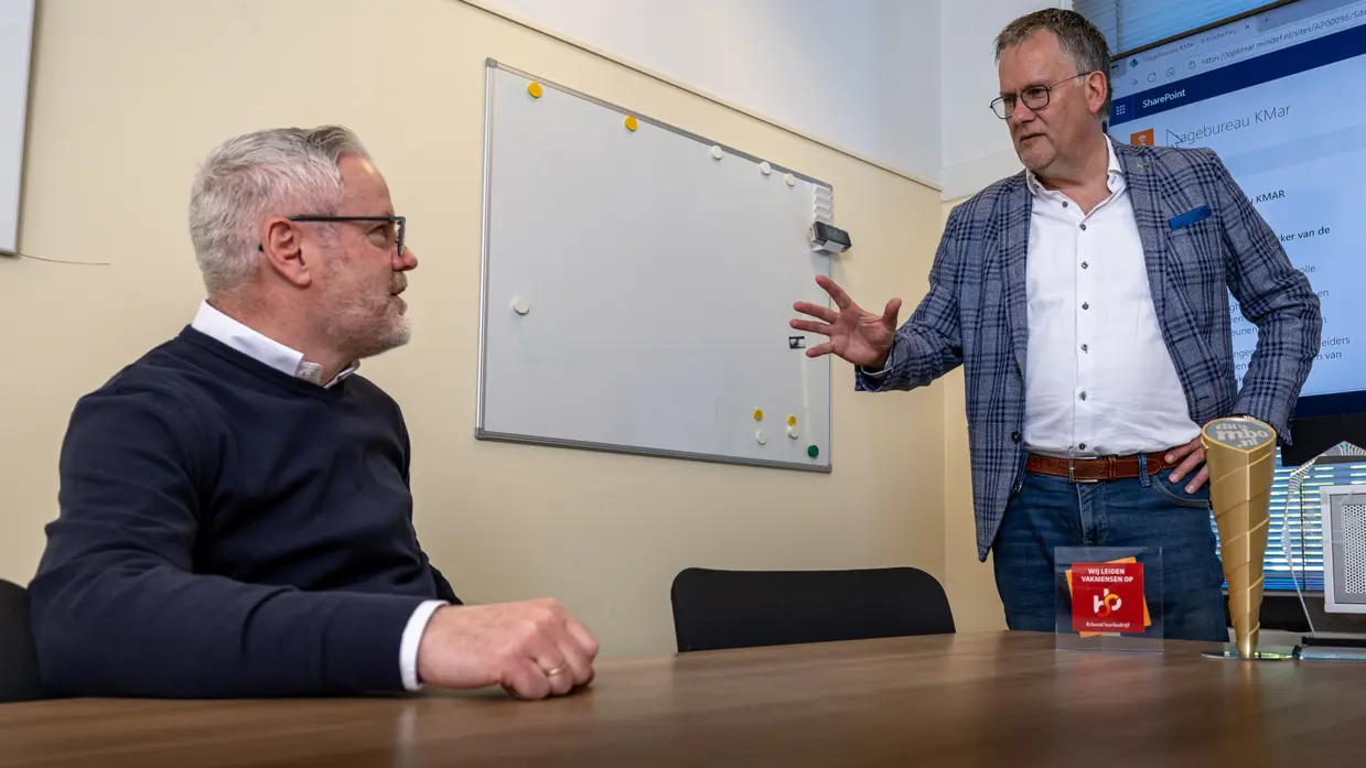 Een man in een blauw, geruit colbert legt handgebaren iets uit aan de zittende man. Beiden dragen een bril.