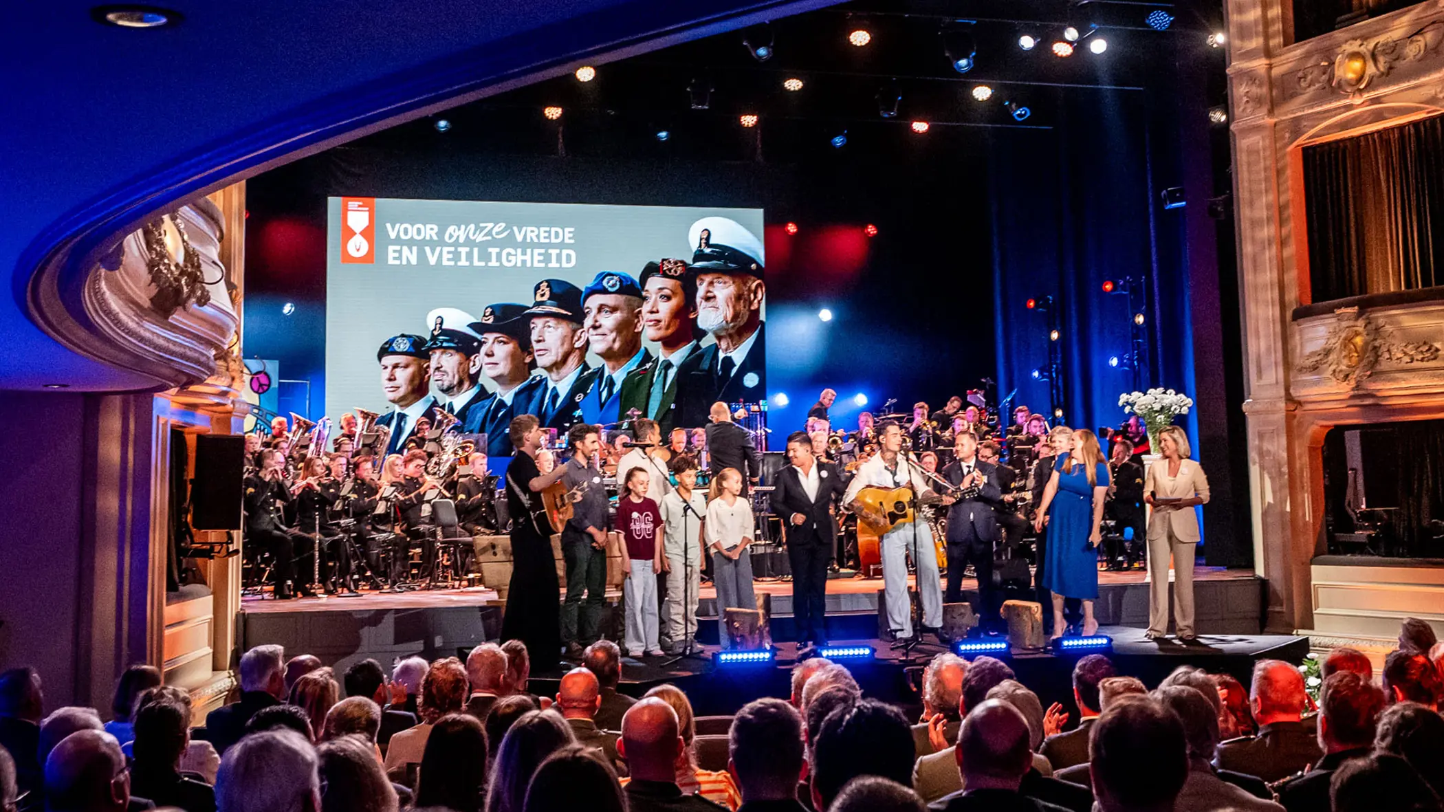 De finale van de ochtend met alle solisten, sprekers, presentatrice en een aantal kinderen op het podium. In de achtergrond is boven het orkest groot een afbeelding van Nederlandse veteranen te zien.