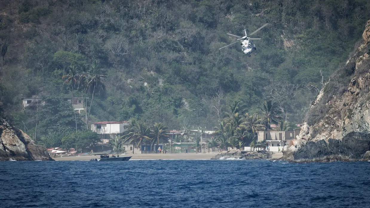 Een helikopter vliegt richting de lens, op de achtergrond groene bergen en wat huisjes.