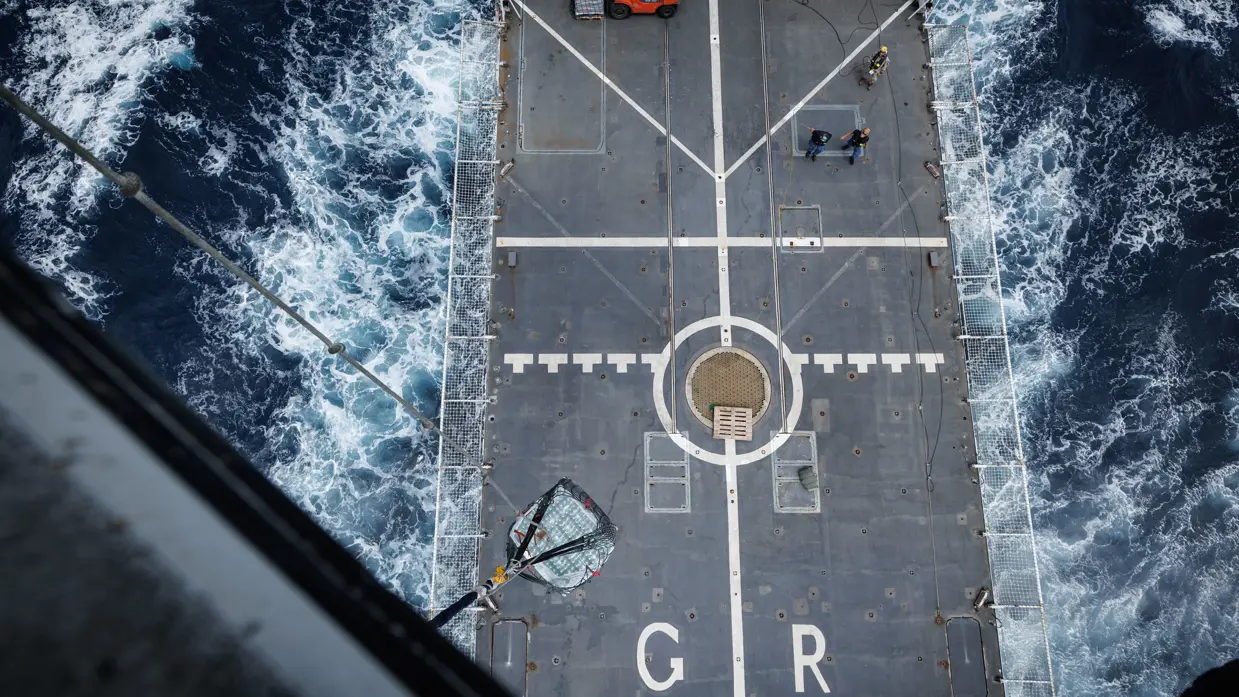 Het grijze helikdek gezien vanuit de helikopter, terwijl het schip vaart.