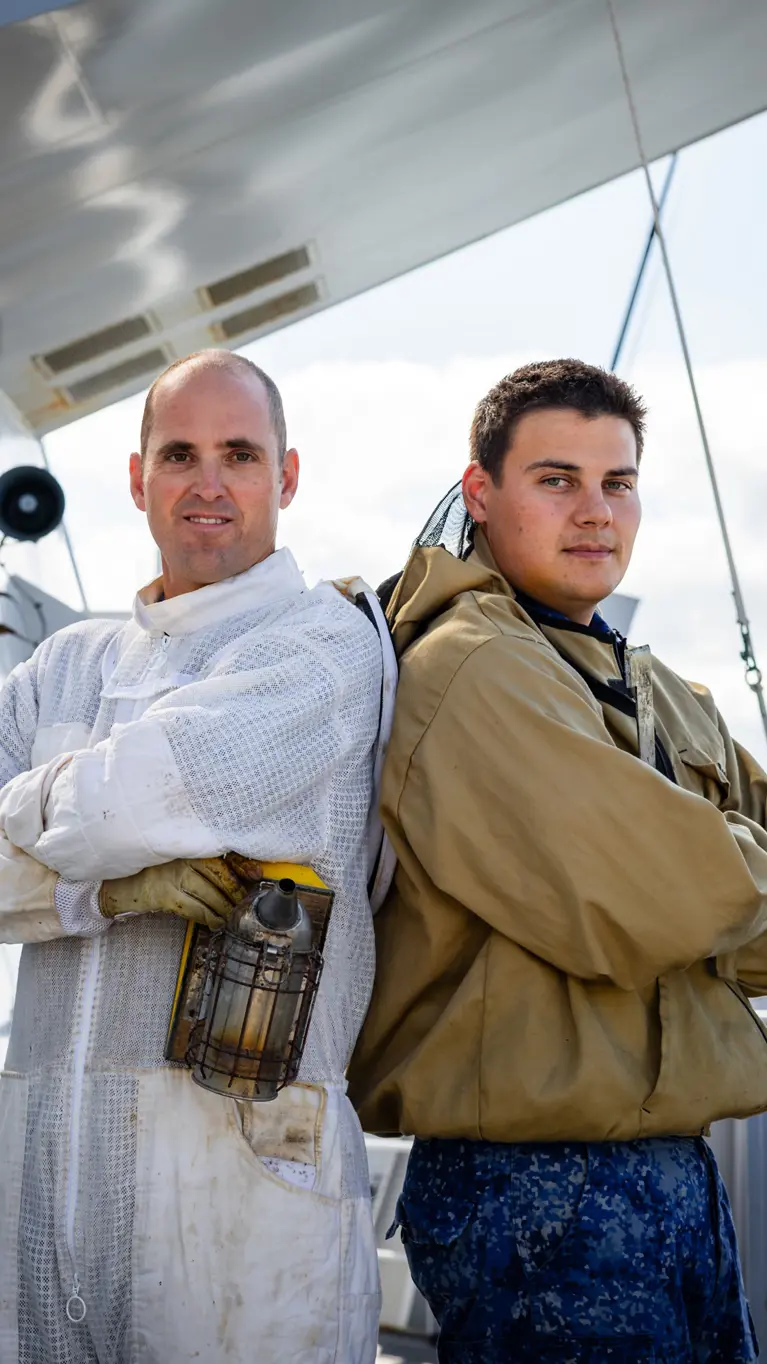 Twee mannen staan in werkpakken op een schip, met pijpen, tanks en kabels onder een heldere lucht.