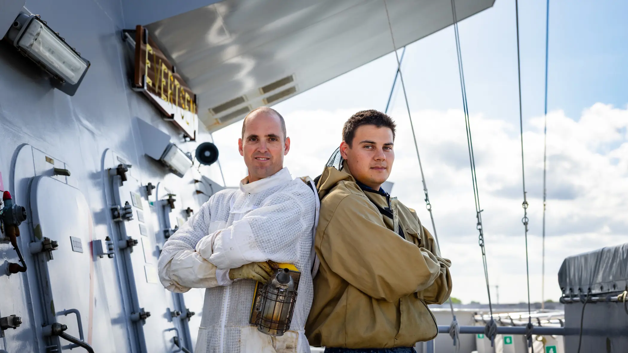 Twee mannen staan in werkpakken op een schip, met pijpen, tanks en kabels onder een heldere lucht.