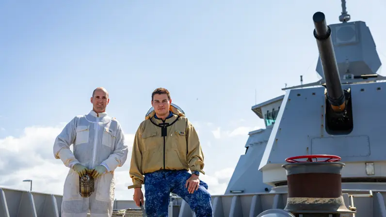 Twee mannen in werkpakken staan op een schip met een grote kanonsloop en een rode kraan op de achtergrond onder een heldere hemel.