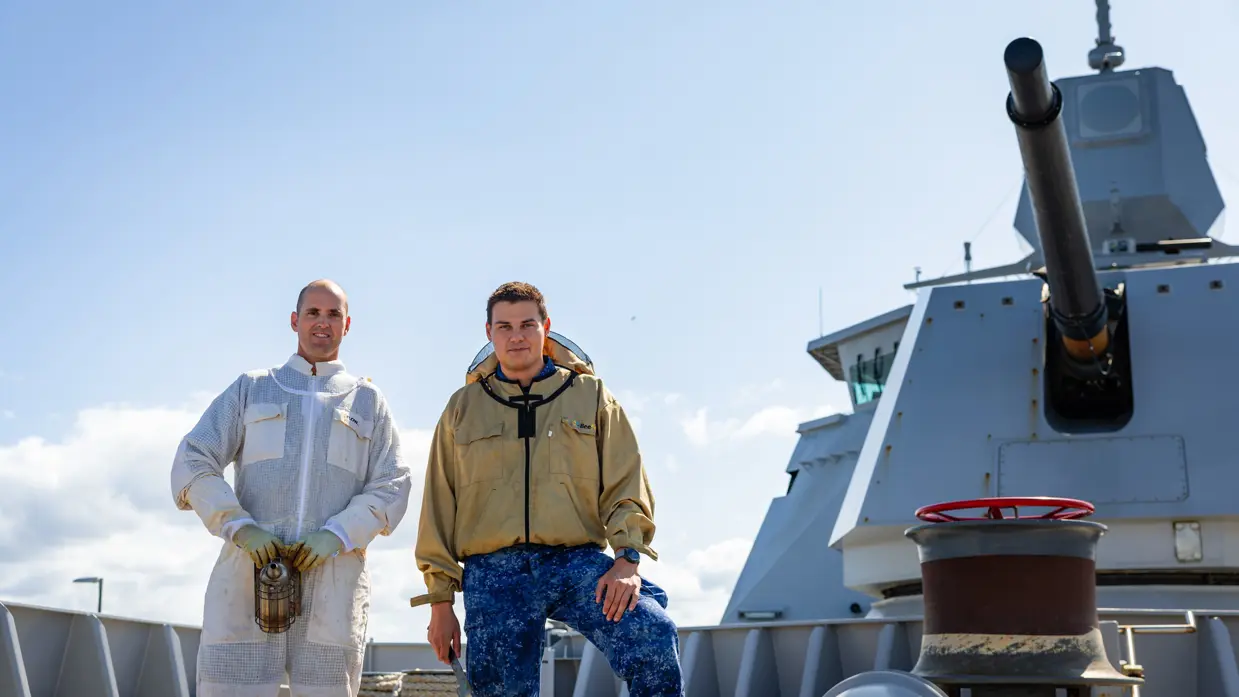 Twee mannen in werkpakken staan op een schip met een grote kanonsloop en een rode kraan op de achtergrond onder een heldere hemel.