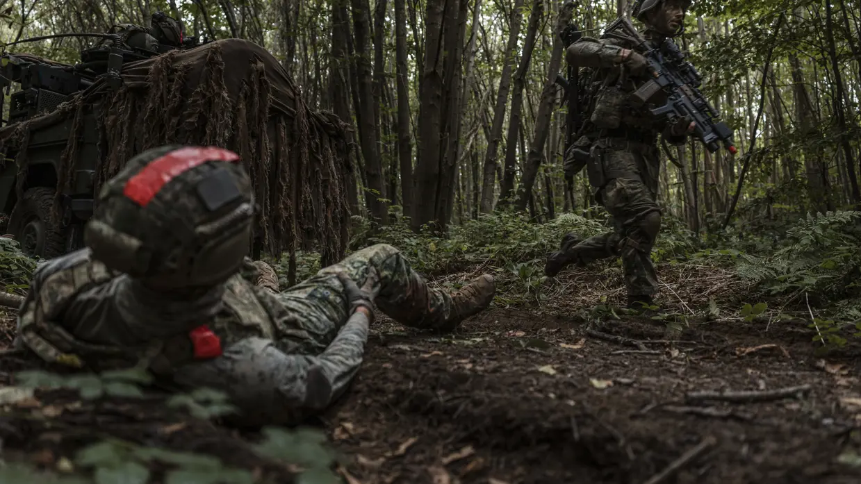 Een militair loopt door het bos, op de voorgrond een gewonde oefenvijand.