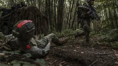 Een militair loopt door het bos, op de voorgrond een gewonde oefenvijand.