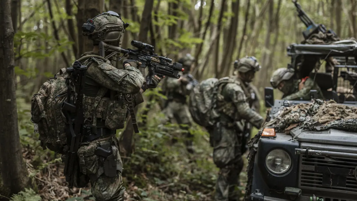 Een militair houdt een collega in de gaten die een voertuig inspecteert.