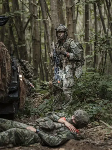 Een militair staat in het bos, op de voorgrond een gewonde oefenvijand.