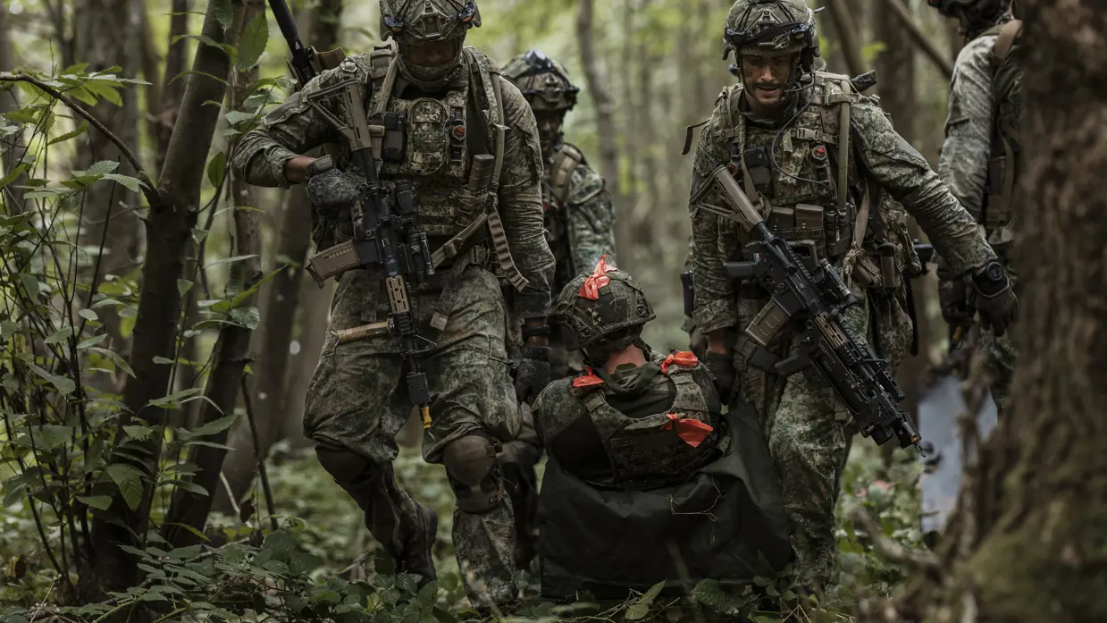 Een groep soldaten sleept een gewonde oefenvijand op een provisorische brancard door het bos.