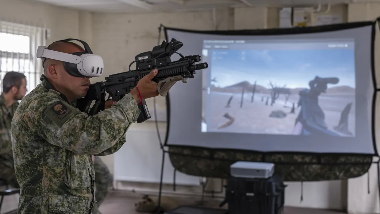 Een militair met een VR-bril voor een scherm waarop hij ‘mee schiet’.