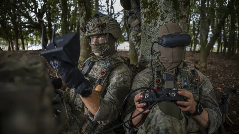 Twee militairen zitten tegen een boom en besturen een drone, eentje met VR-bril.