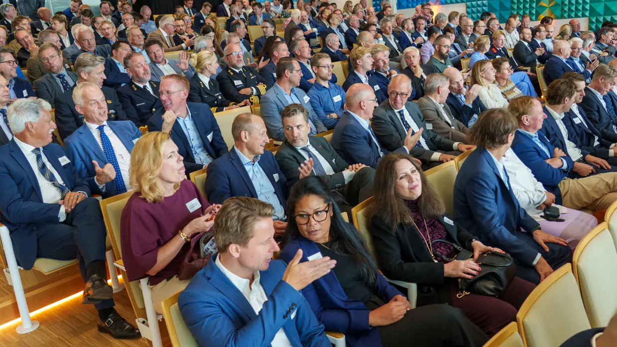 Een militair speelt op een keyboard met achter zich een groot projectiescherm en een commandeur spreekt een zaal toe met achter zich een groot projectiescherm.