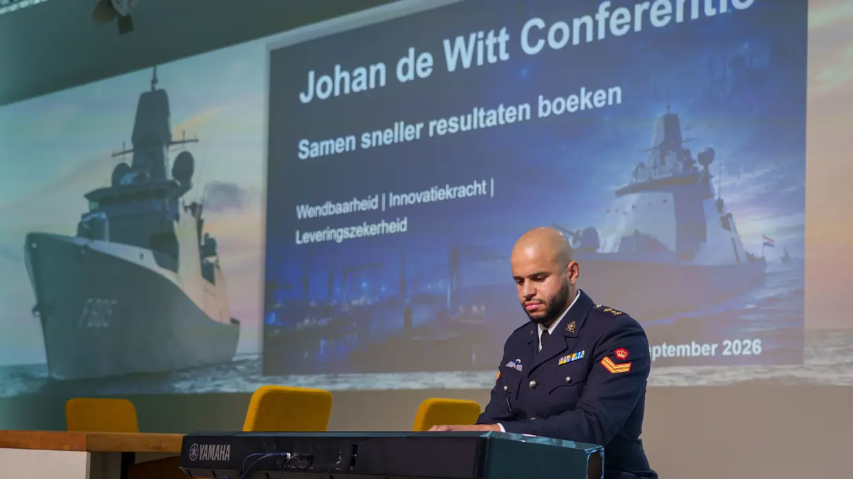 Een militair speelt op een keyboard met achter zich een groot projectiescherm en een commandeur spreekt een zaal toe met achter zich een groot projectiescherm.