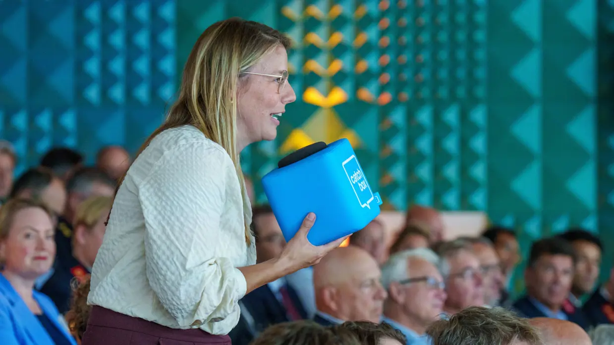Een bebrilde vrouw spreekt in een volle zaal door een catchbox. 