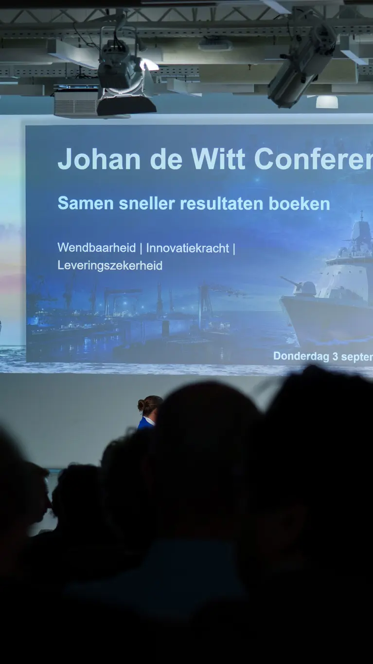Een blik vanuit het publiek op het podium en grote projectiescherm. 