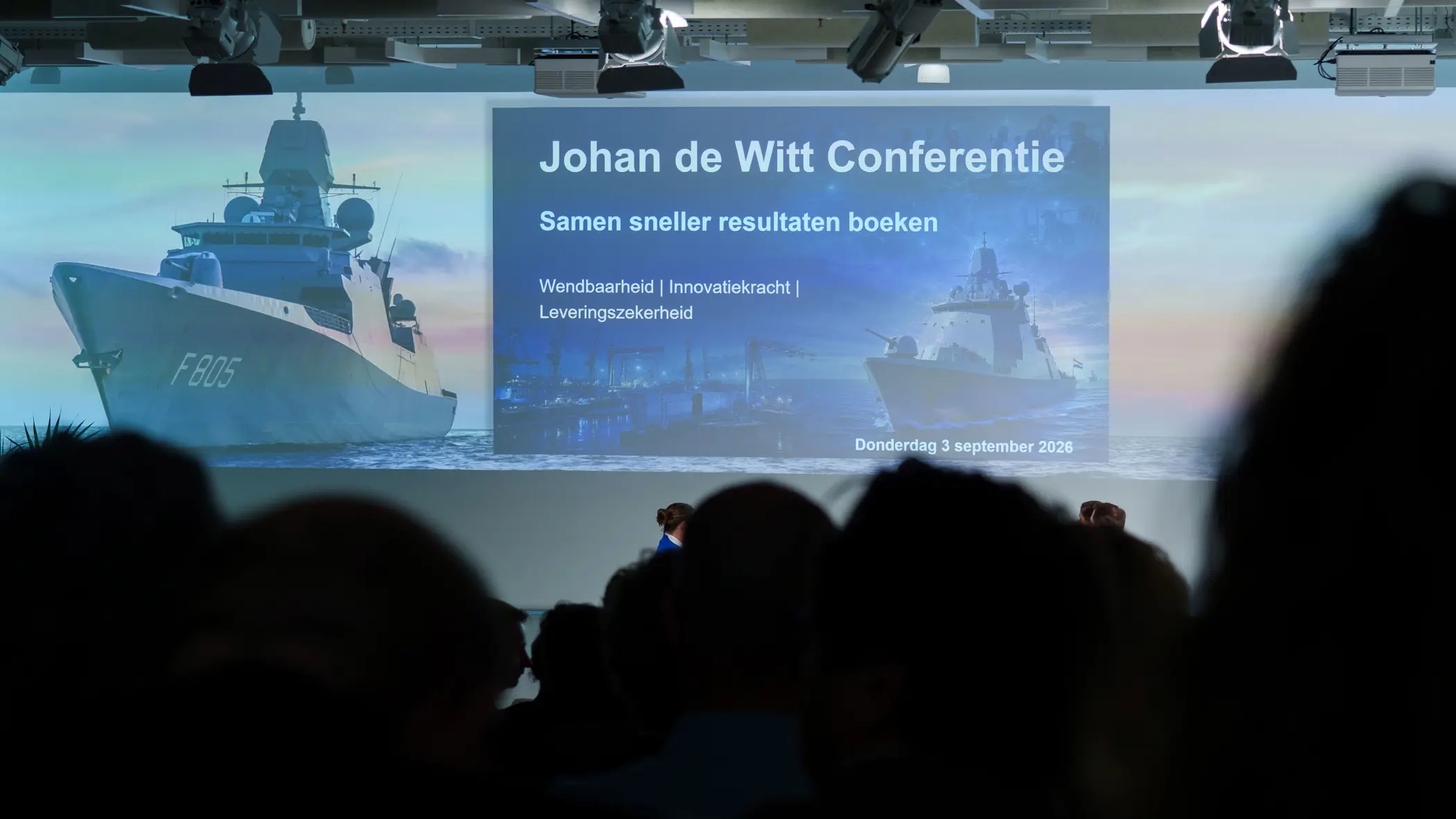 Een blik vanuit het publiek op het podium en grote projectiescherm. 