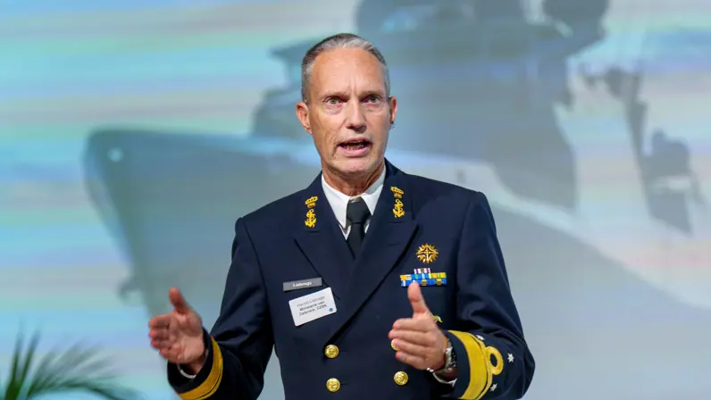 Commandant Zeestrijdkrachten spreekt voor een afbeelding van groot marineschip op een projectiescherm en de conferentiezaal met toehoordes en links de admiraal zelf.