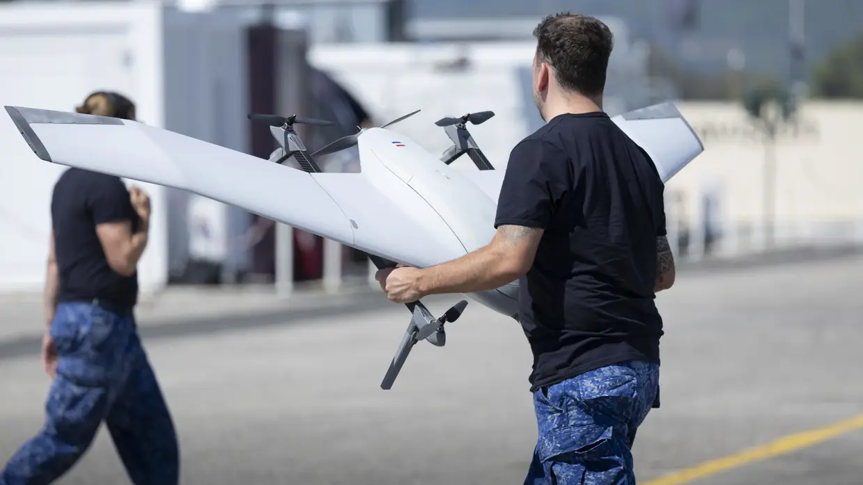 Een man gehuld in de broek van het boordtenue en een donker T-shirt houdt een grijze drone vast. 