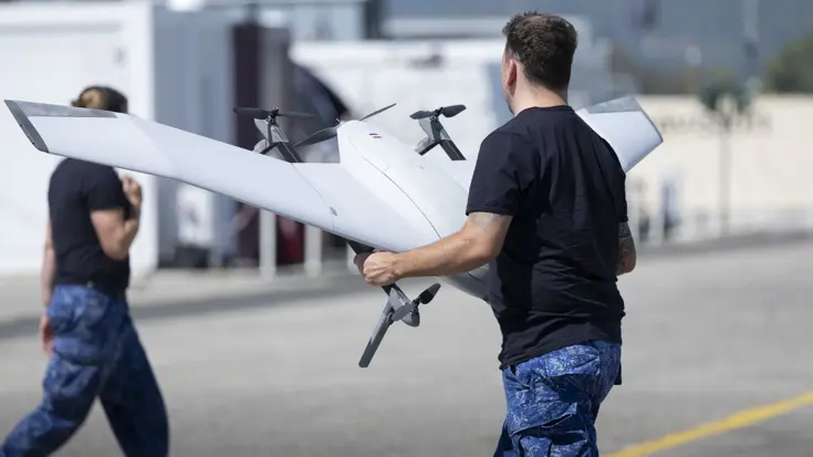 Een man gehuld in de broek van het boordtenue en een donker T-shirt houdt een grijze drone vast. 