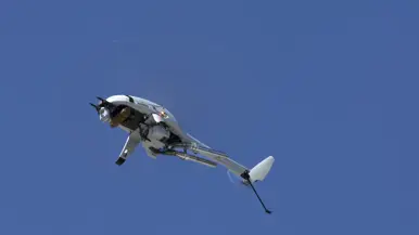 Een grijze helikopterdrone vliegt onder een strakblauwe hemel.