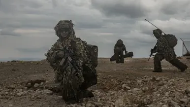 Een militair in een speciaal camouflagepak zit geknield en scant de omgeving terwijl achter hem collega’s hun stelling innemen.