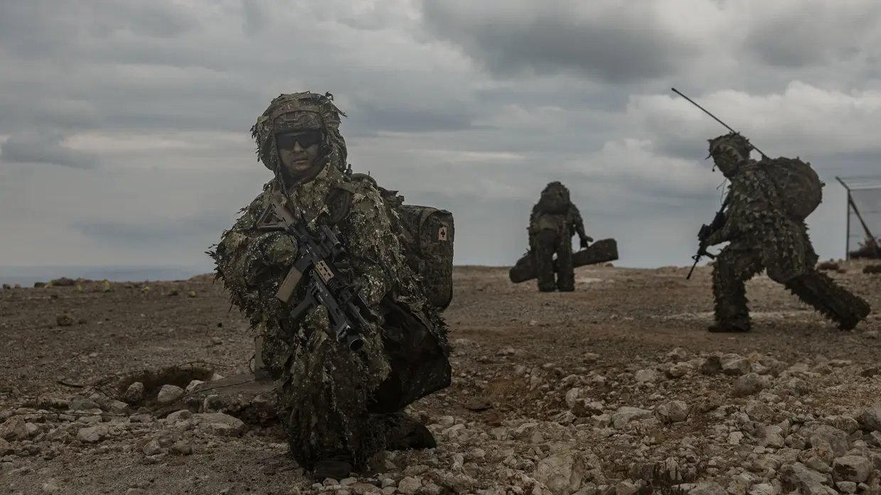 Een militair in een speciaal camouflagepak zit geknield en scant de omgeving terwijl achter hem collega’s hun stelling innemen.