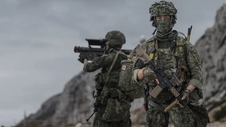 Een militair in vol ornaat, met een buff voor zijn gezicht poseert met wapen voor de camera. Op de achtergrond richt een militair een Stinger op de lucht.