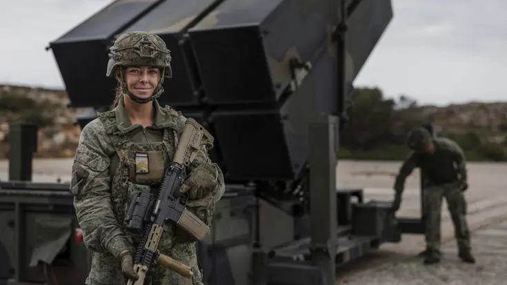 Een vrouwelijke militair in vol ornaat met geweer poseert voor een NASAMS-luchtverdedigingssysteem.