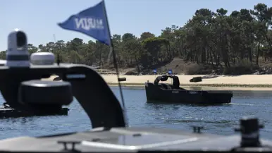 Een onbemand bootje vaart over kalm water. Daarachter de kustlijn met zand en bomen.