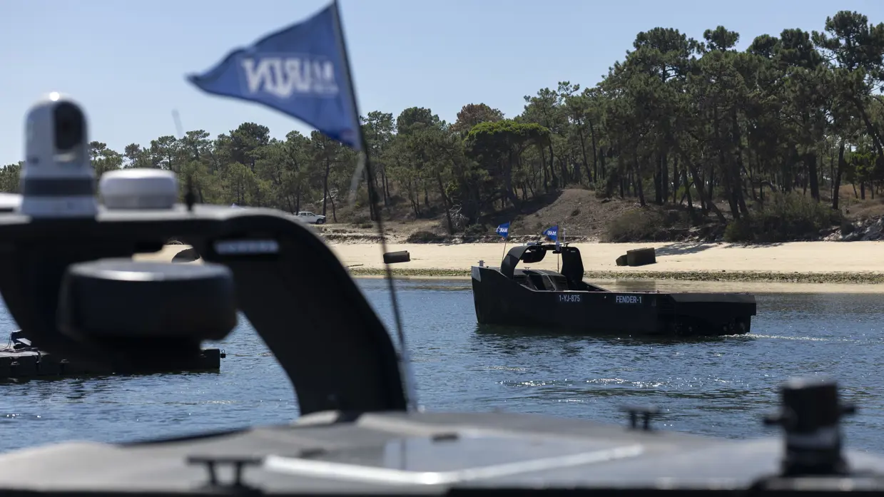 Een onbemand bootje vaart over kalm water. Daarachter de kustlijn met zand en bomen.