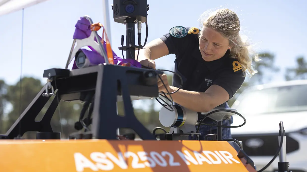 Een blonde vrouw met donkerblauw T-shirt sleutelt aan een drone met een oranje zijkant.