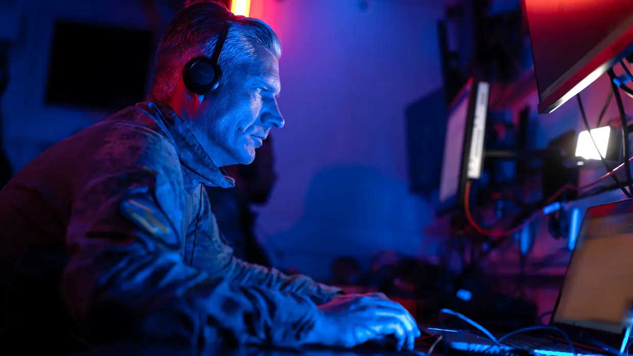 Portretfoto van kapitein Wibo in een donkere werkcontainer, hij zit achter enkele computerschermen en heeft een headset op.