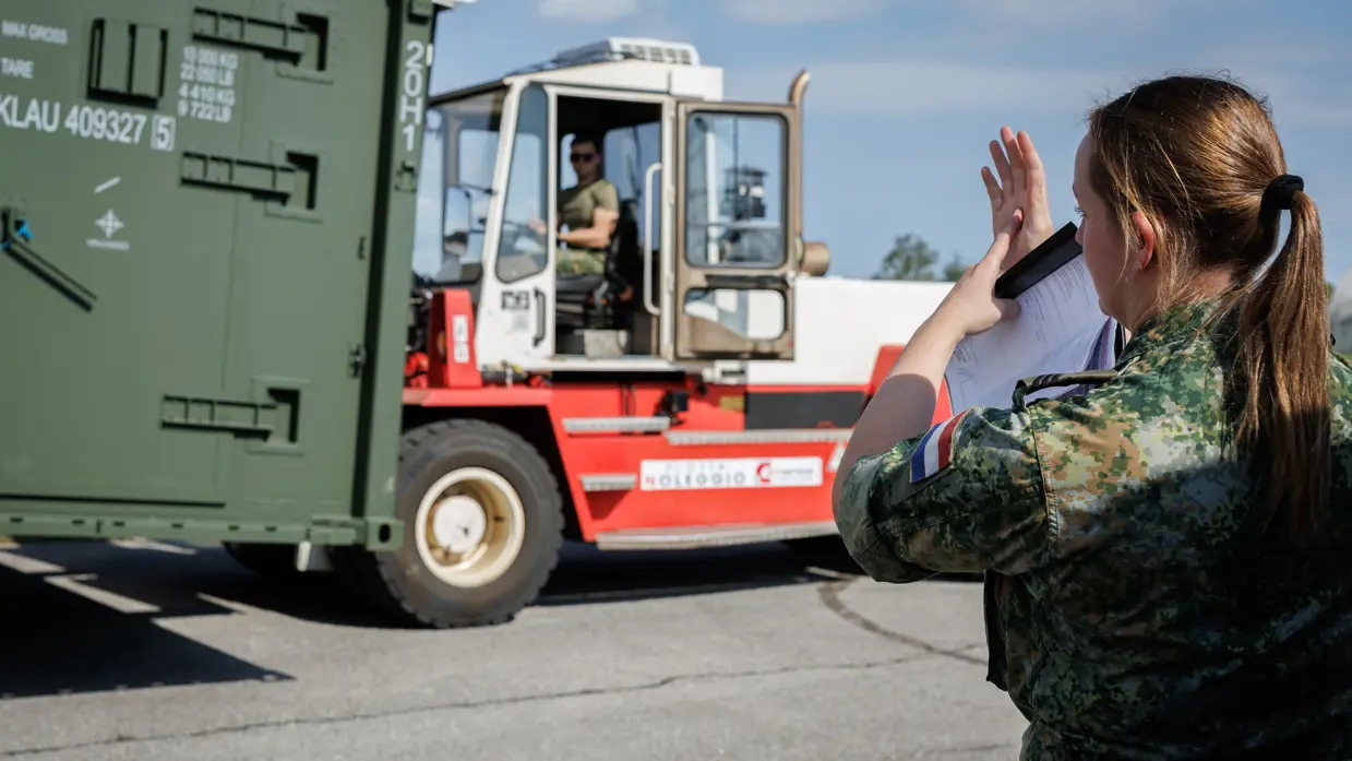 Vrouwelijke militair met boekje in de hand maakt handgebaren naar een heftruckchauffeur die een container vervoert.
