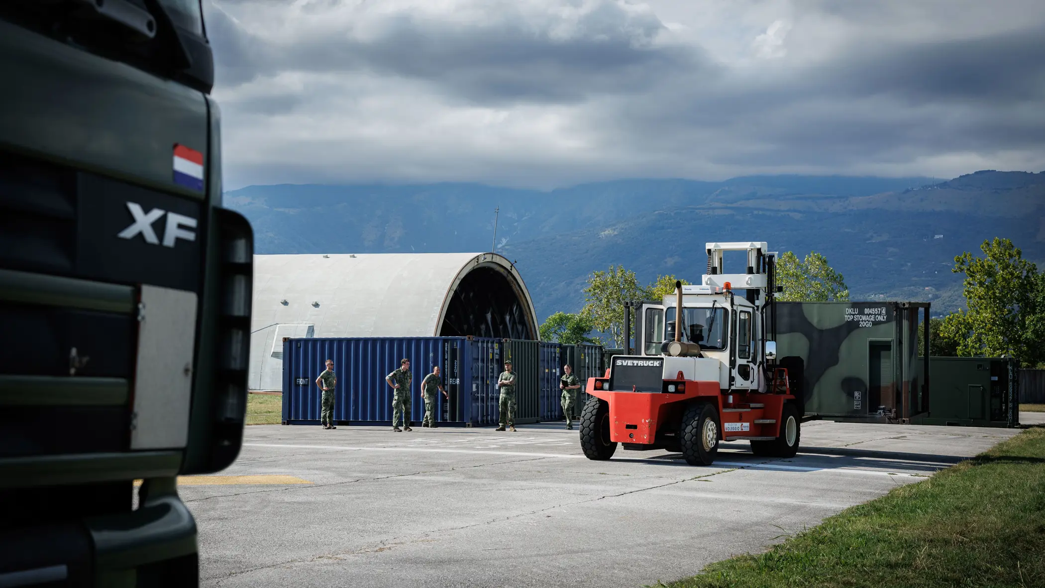 Een heftruck met een container rijdt over een betonnen ondergrond tussen gras gelegen. Links meerdere containers en een oude vliegtuigshelter. Op de achtergrond bergen.